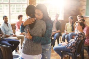 Two women hugging in a room with people sitting around.